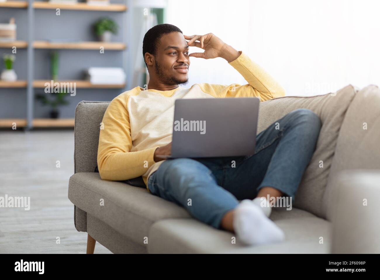 Un ragazzo nero sognante con un computer portatile appoggiato sul divano a casa Foto Stock