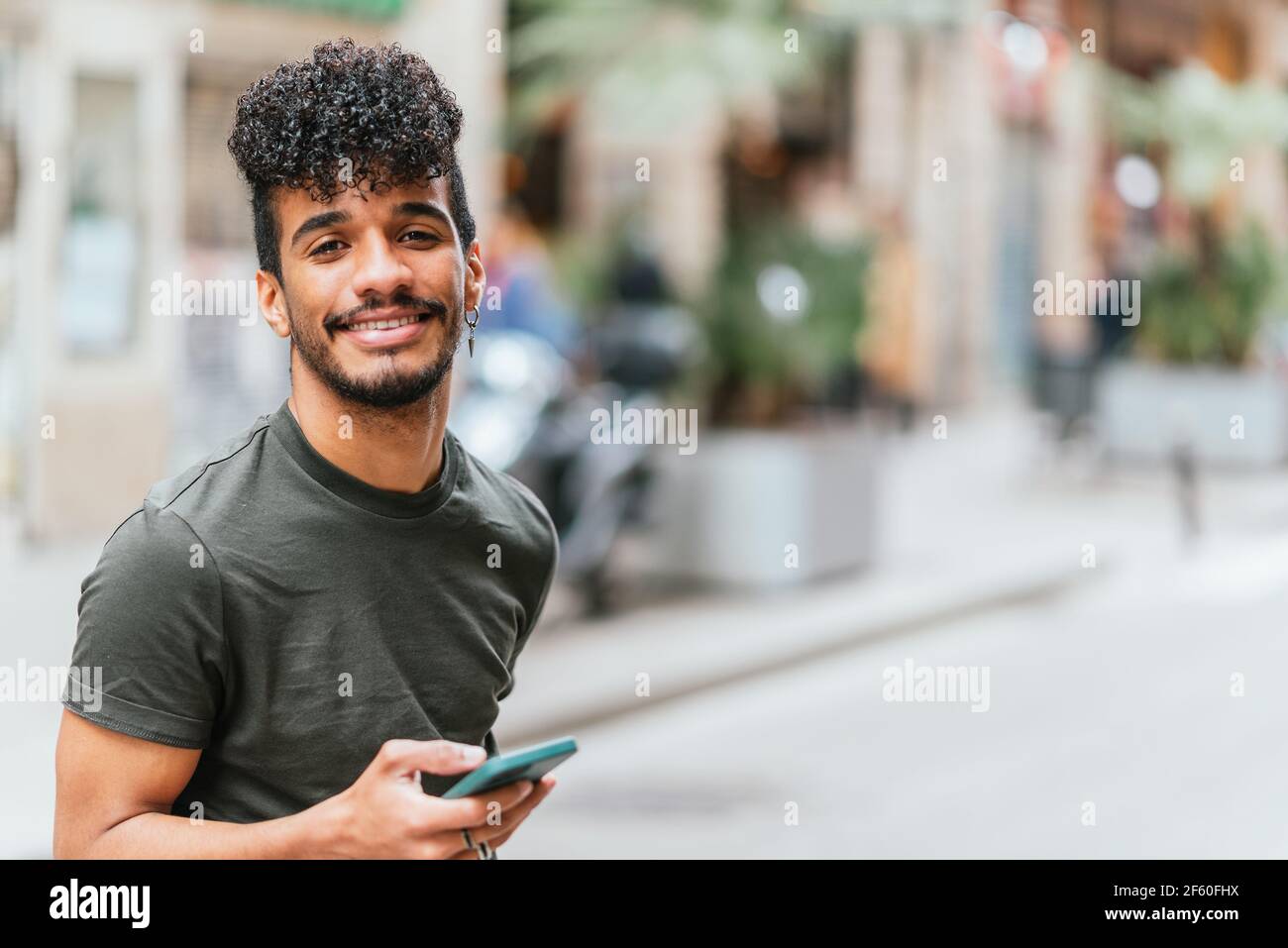 ritratto di un giovane e attraente latino-americano guardando la macchina fotografica e sorridendo. Sta tenendo un telefono cellulare blu. È in strada a Barcellona. Foto Stock