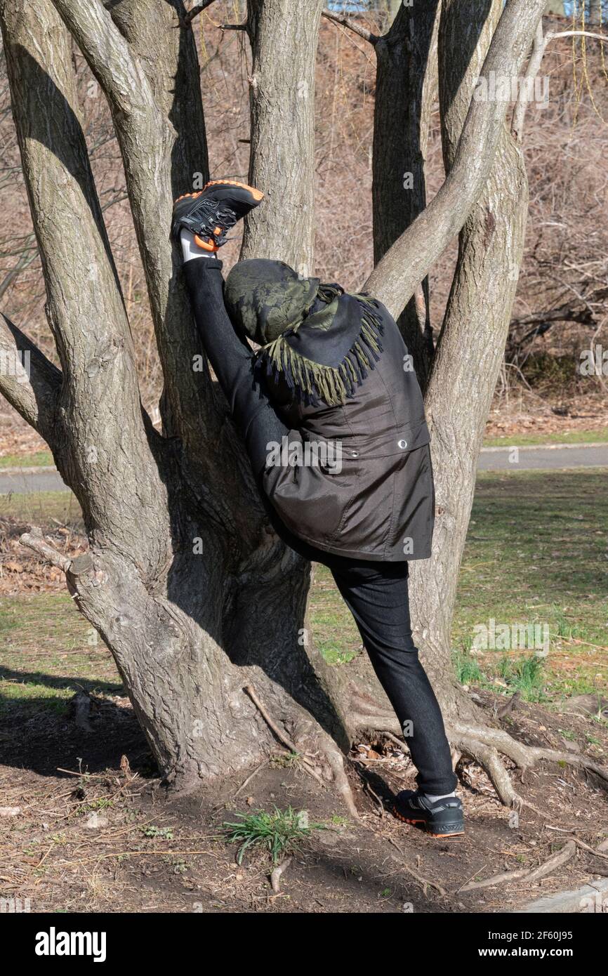 una donna estremamente flessibile allunga la gamba di un piede contro un albero. A Kissena Park, Flushing, New York. Foto Stock