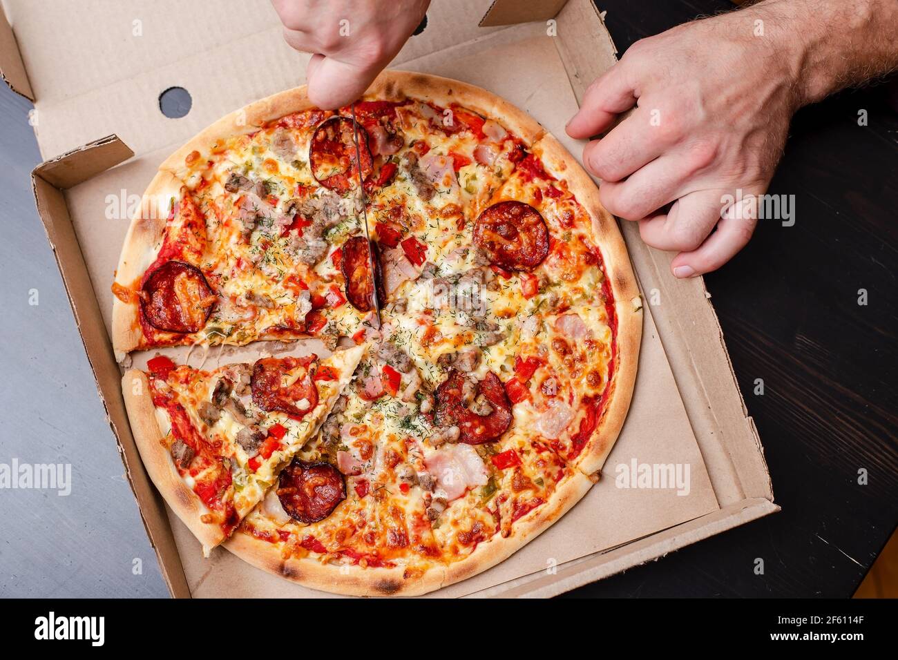 La mano dell'uomo sta tagliando una pizza appetitosa a fette, in una scatola, su un tavolo nero. Vista dall'alto. Foto Stock