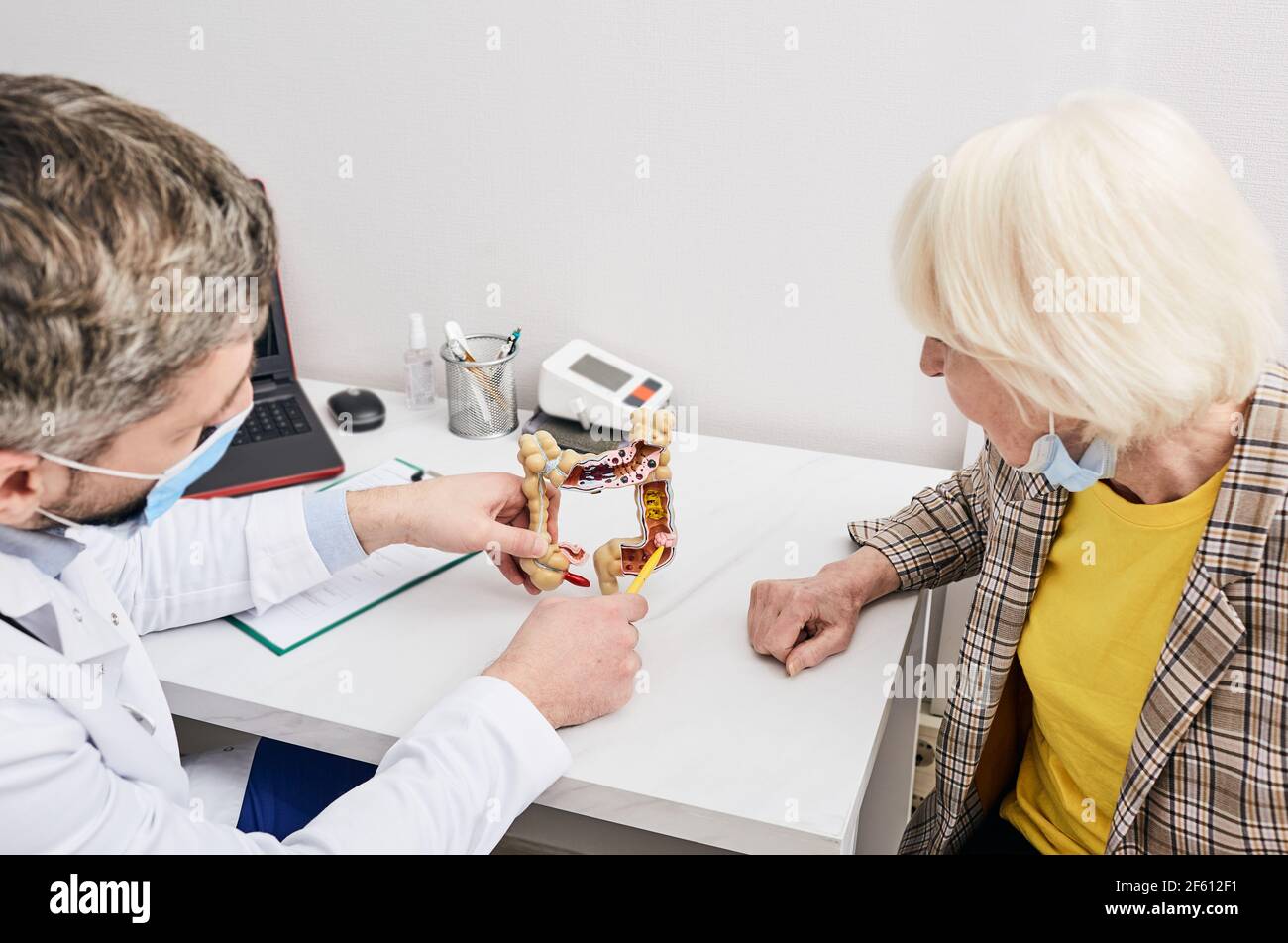 Cancro all'intestino. medico che indossa una maschera medica mostra a donna anziana intestini modello anatomico durante una visita alla clinica Foto Stock