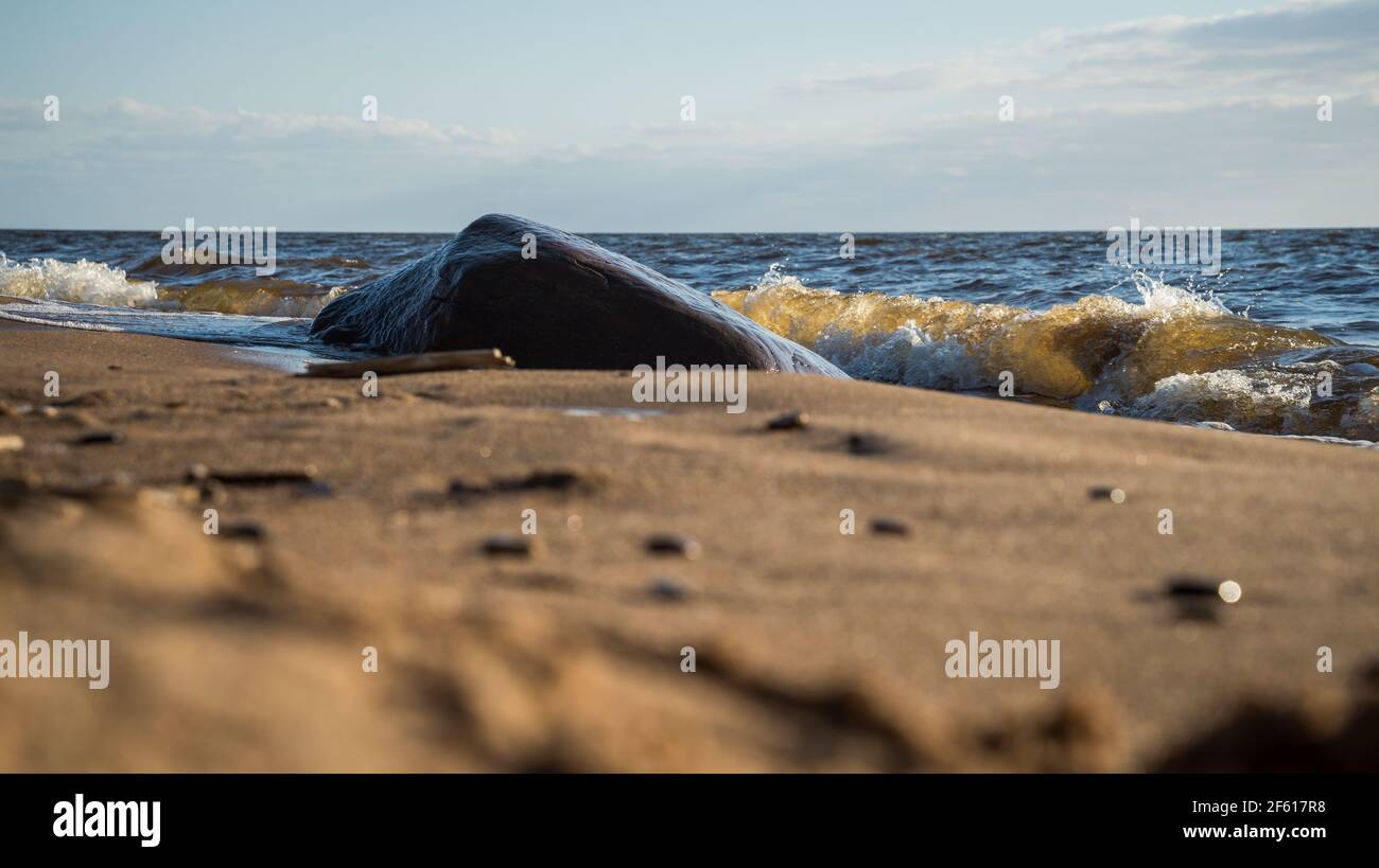 Bella grande masso marrone sulla spiaggia di mare bagnata da belle onde d'acqua di mare. Il primo piano è sfocato con piccoli ciottoli di mare. Bellissima Foto Stock