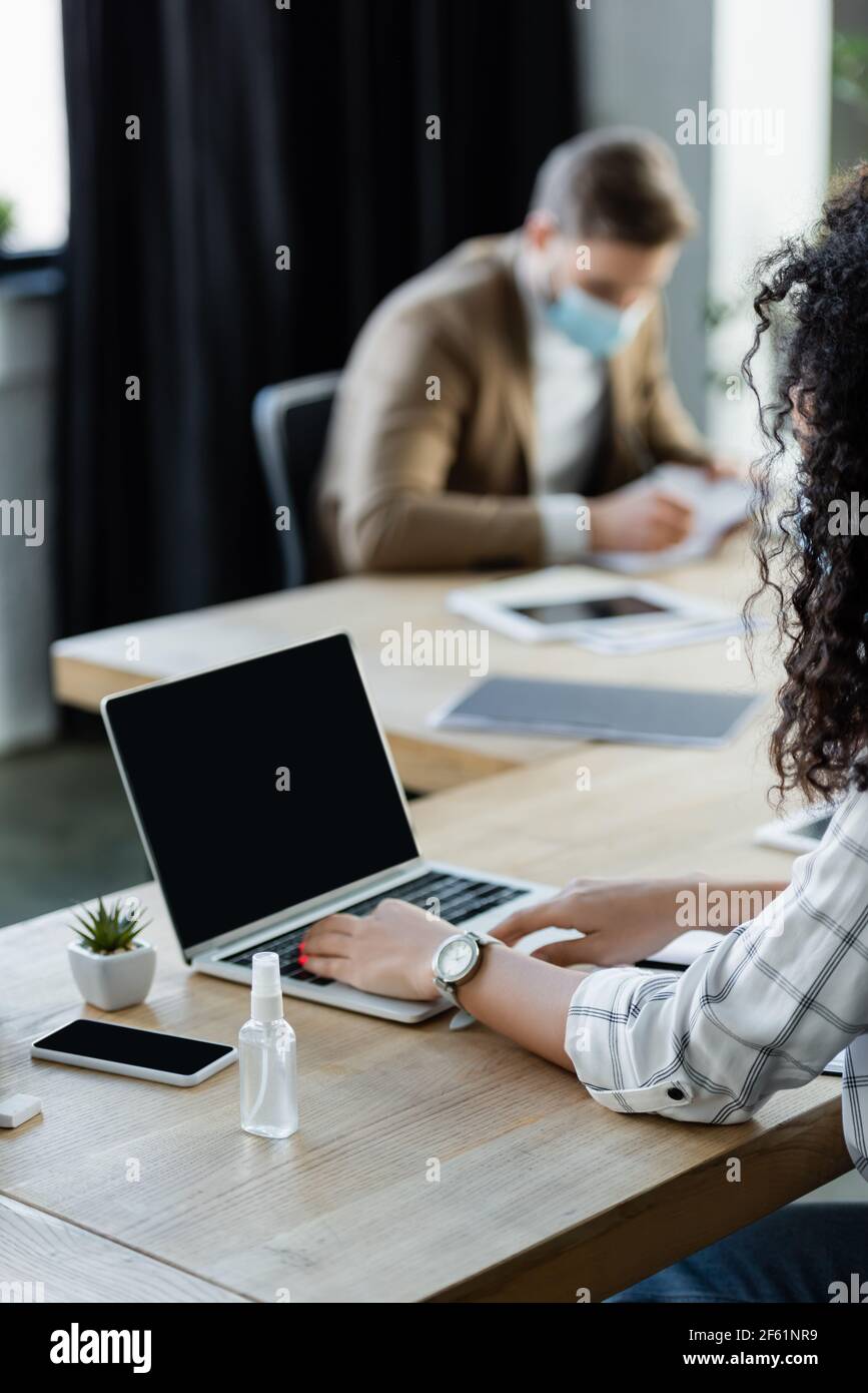 donna d'affari amerian africana che scrive sul laptop vicino manager in medicina maschera di lavoro su sfondo sfocato Foto Stock