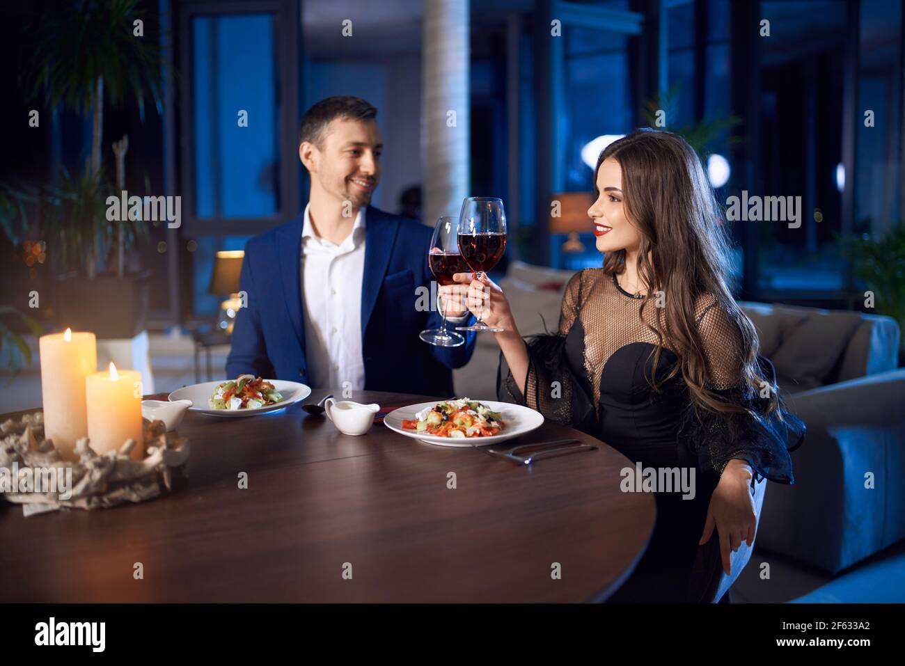 Giovane uomo e donna seduti a tavola, bevendo vino rosso e mangiando insalata fresca. Una coppia elegante che fa un appuntamento romantico mentre si soggiorna a casa. Foto Stock