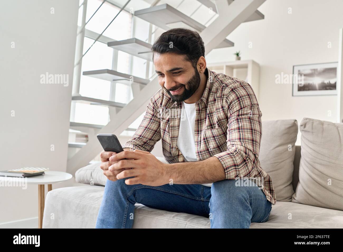 Felice giovane indiano che usa lo smartphone seduto sul divano a casa. Foto Stock