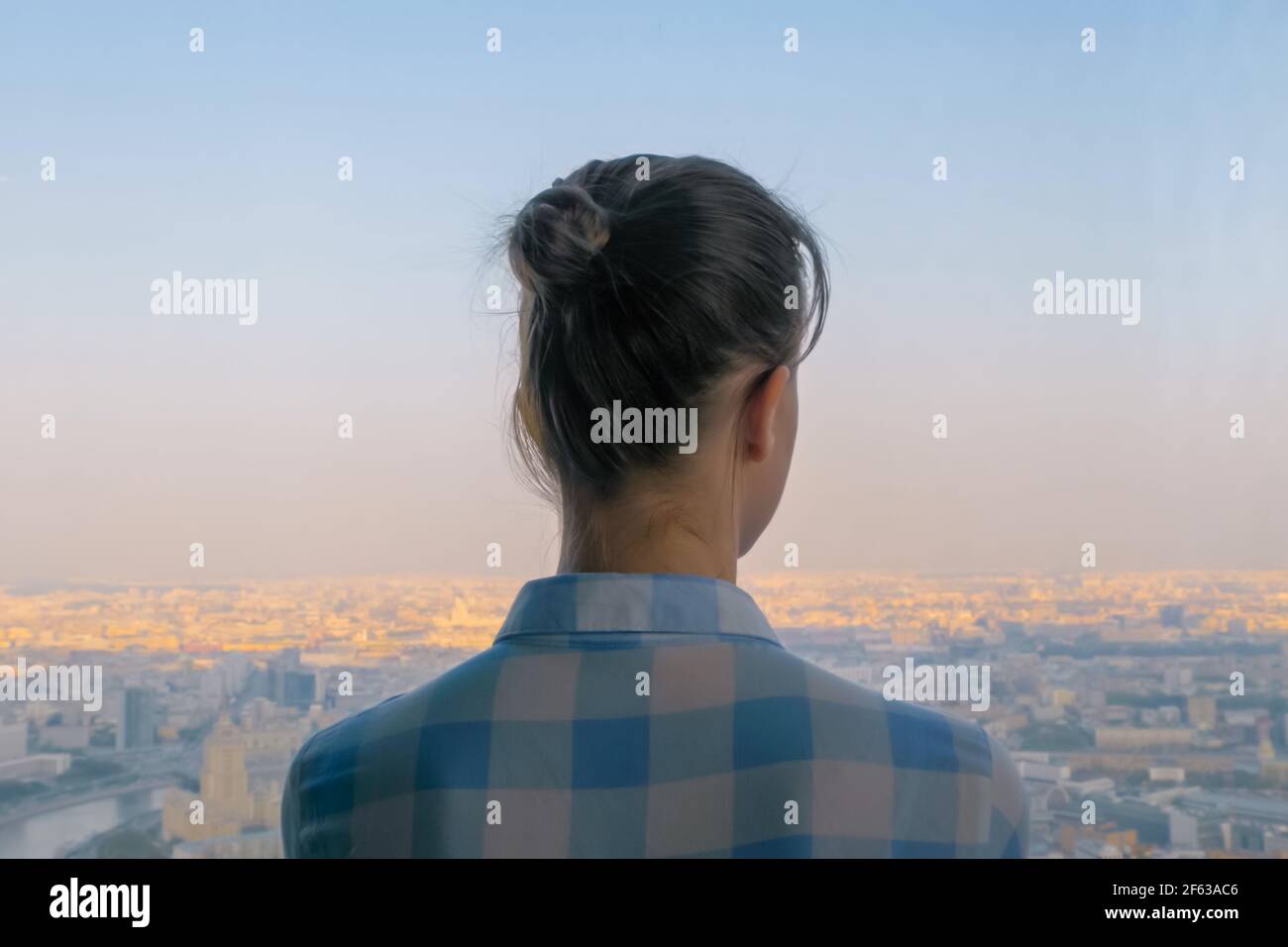 Vista posteriore di una donna penosa che guarda il paesaggio urbano attraverso la finestra di grattacieli Foto Stock