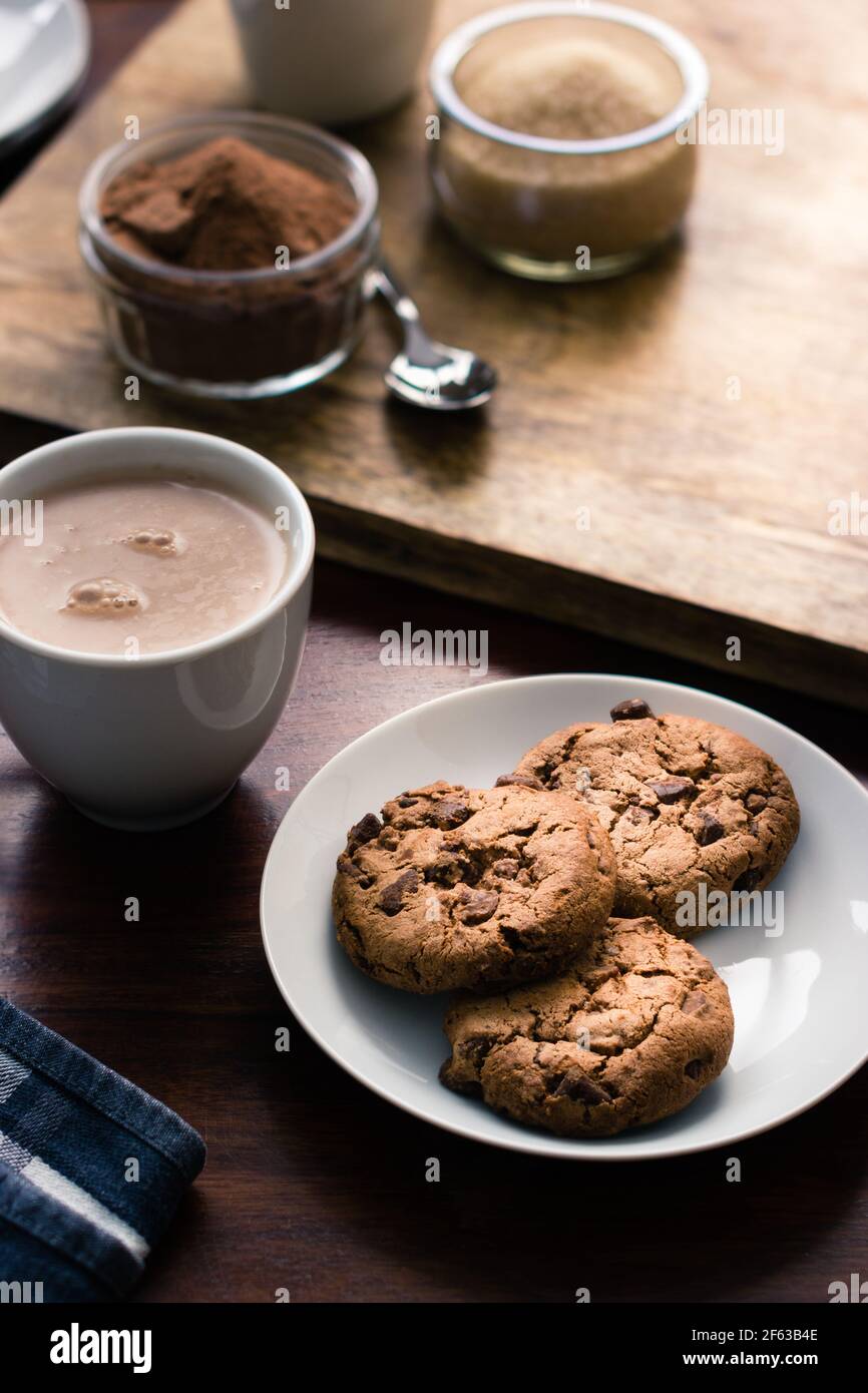 Pila di biscotti su un piatto accanto a una tazza di bere cioccolato prima di zucchero e barattoli di cacao in polvere Foto Stock