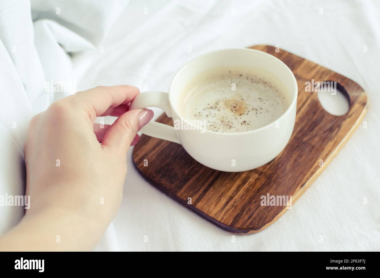 Una tazza di caffè in mano femminile nel letto su una coltre bianca. Moman godendo del comfort a casa accogliente atmosfera. Buona mattina. POV. Foto Stock