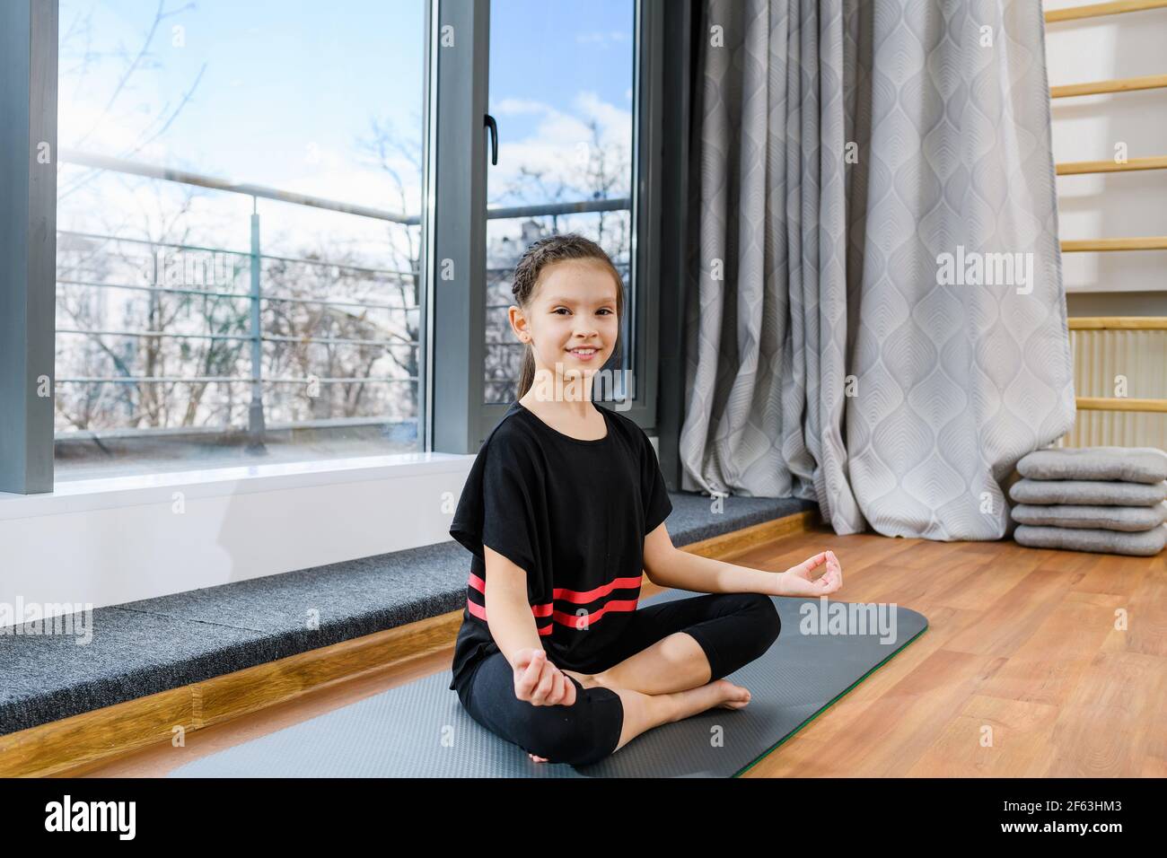 Pre ginnast sorridente bambina seduta su tappeto sportivo in lotus yoga posa vicino alla finestra panoramica, infanzia sana Foto Stock