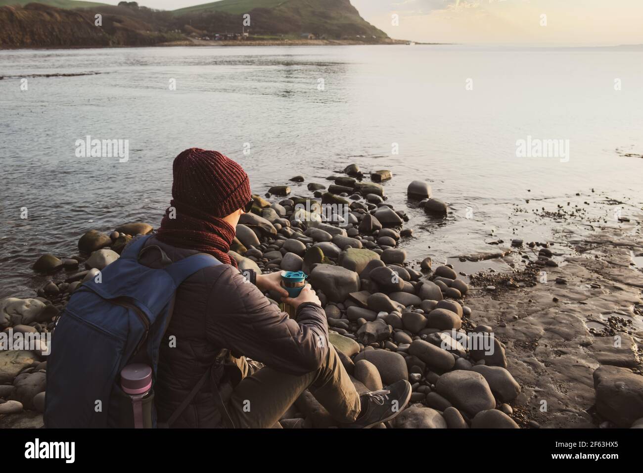 Un giovane in abiti caldi seduto sulla pietra, bevendo tè caldo o caffè da thermos riutilizzabile tazza e godendo vista mare. Rilassati durante una passeggiata Foto Stock