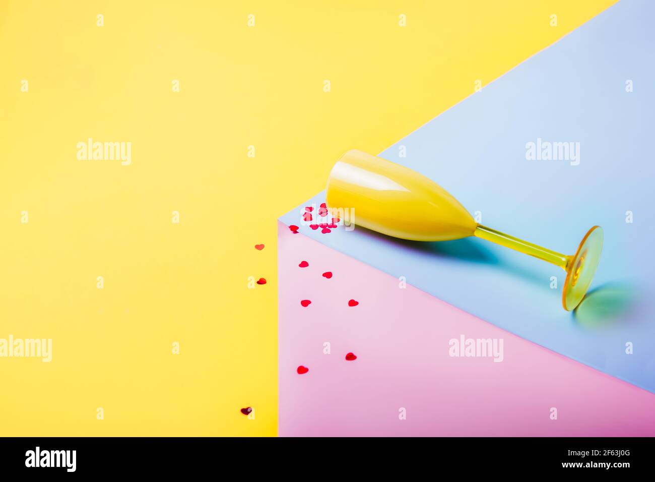 Confetti rossi a forma di cuore cadenti dal bicchiere di vino giallo adagiati sul podio rosa e blu sullo sfondo giallo. Concetto di amore creativo. San Valentino Foto Stock