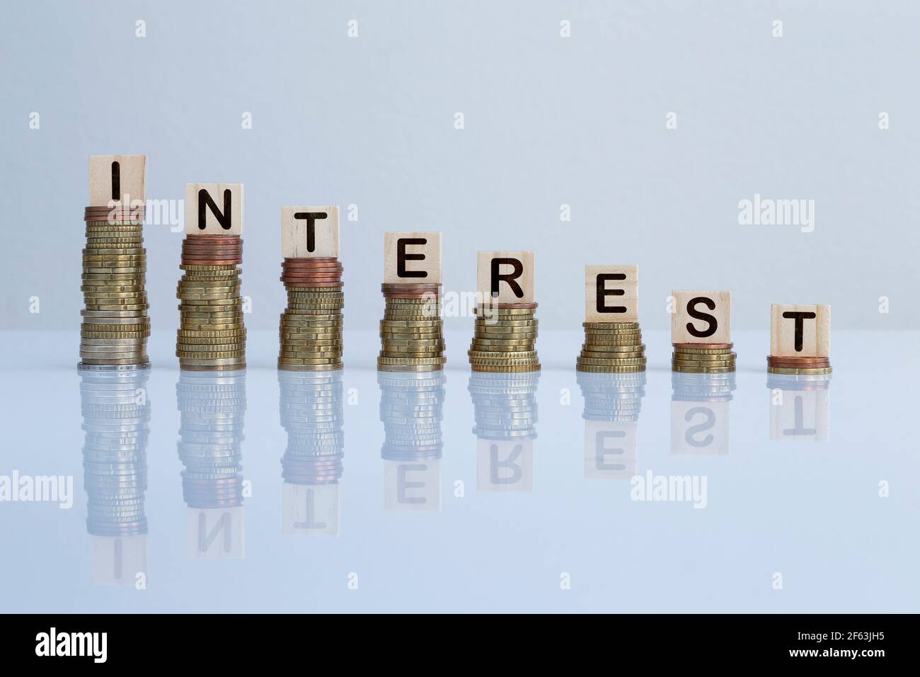 Parola 'INTERESSE' su blocchi di legno in cima a pile discendenti di monete su sfondo grigio. Foto concettuale di economia, affari, finanza e banche Foto Stock