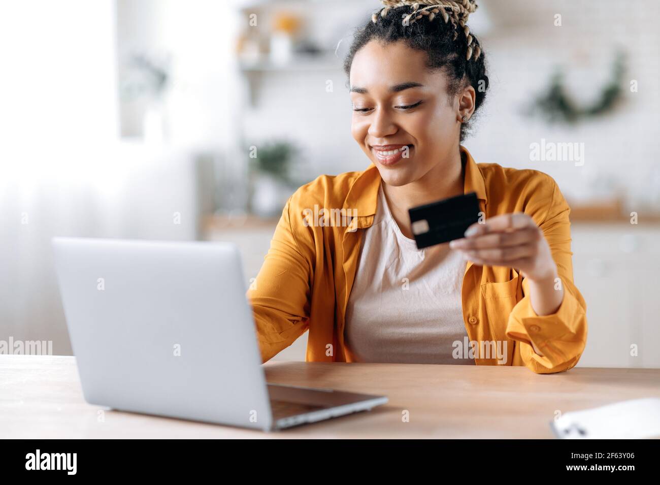 Felice giovane afroamericana donna elegantemente vestito, pagando per gli acquisti online, usa un laptop, tenendo una carta bancaria in mano, digitando le informazioni della carta di credito sulla tastiera e sorridendo Foto Stock