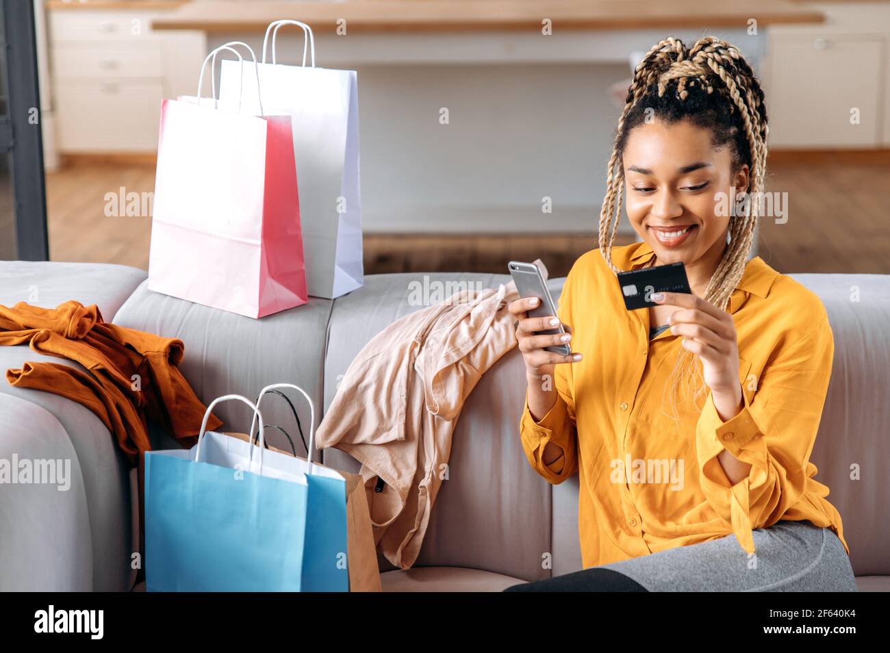 Giovane donna afroamericana attraente di successo seduta sul divano a casa, utilizzando smartphone e carta bancaria per i pagamenti online, molte borse di carta vicino, inserire il numero di carta, sorridendo Foto Stock