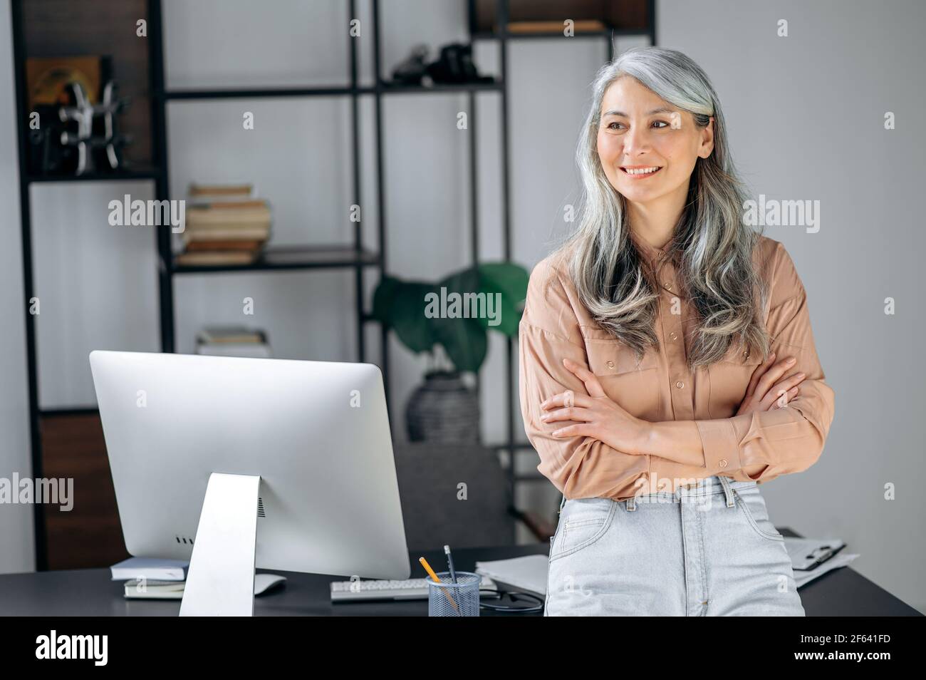 Ritratto di una donna asiatica con capelli grigi abbastanza influente, leader di affari, manager, ceo si trova in un ufficio moderno, elegantemente vestito, braccia incrociate, guardando al fianco, sorridente amichevole Foto Stock