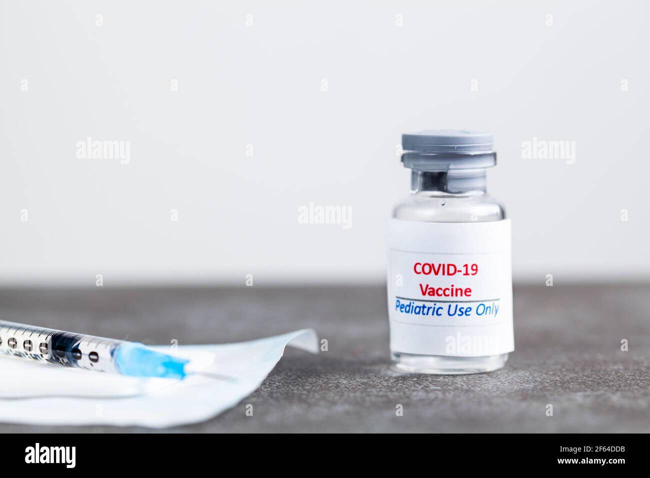 Immagine concettuale che mostra il vaccino COVID 19 progettato per l'uso pediatrico solo in un flaconcino di vetro con un ago e una siringa riempito con il vaccino prima della cenere Foto Stock