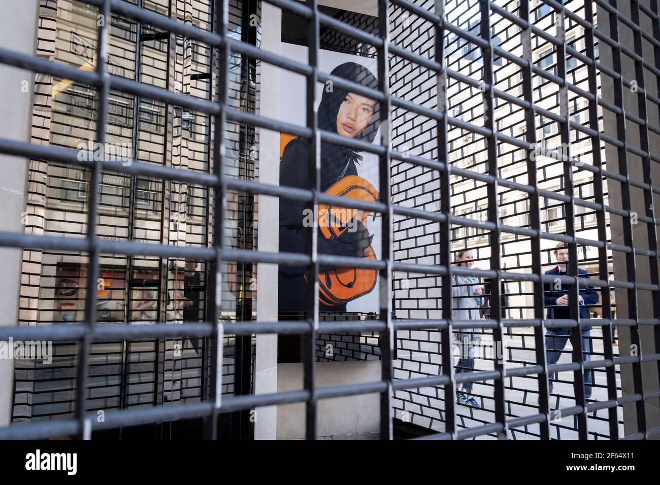 Il volto di un modello maschile si affaccia attraverso la griglia dei cancelli di sicurezza, all'esterno del ramo chiuso di New Bond Street del rivenditore di moda Burberry, durante il terzo blocco della pandemia di Coronavirus, il 29 marzo 2021, a Londra, Inghilterra. Foto Stock