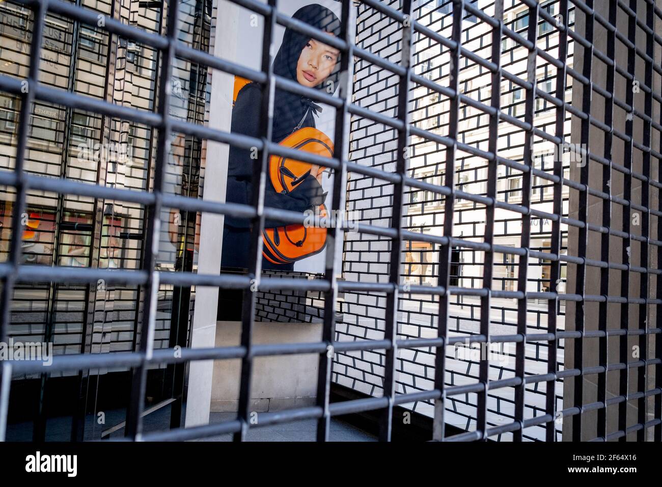 Il volto di un modello maschile si affaccia attraverso la griglia dei cancelli di sicurezza, all'esterno del ramo chiuso di New Bond Street del rivenditore di moda Burberry, durante il terzo blocco della pandemia di Coronavirus, il 29 marzo 2021, a Londra, Inghilterra. Foto Stock