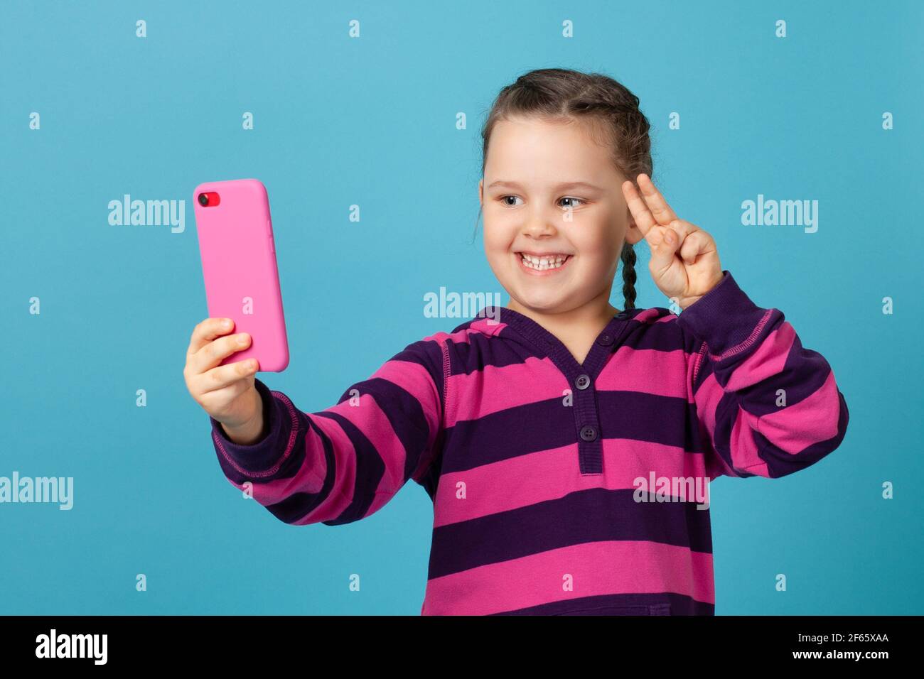 bambina sorridente e scattante selfie, facendo una telefonata su un telefono rosa e mostrando un segno di vittoria con le dita, isolato su uno sfondo blu Foto Stock