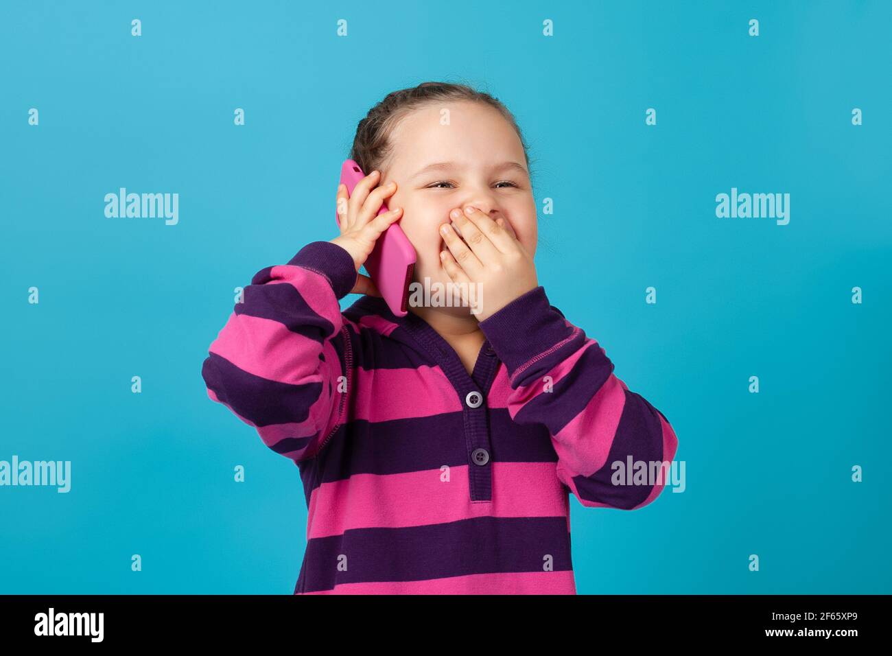 primo piano ritratto di felice ragazza con pigtail che fa una telefonata e che copre la sua bocca con la mano di ridere, isolato su uno sfondo blu Foto Stock