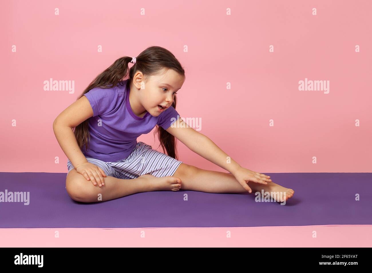 ritratto a tutta lunghezza di un bambino prescolare che fa ginnastica e fa curve laterali mentre si siede su un tappeto di sport viola, isolato su uno sfondo rosa Foto Stock