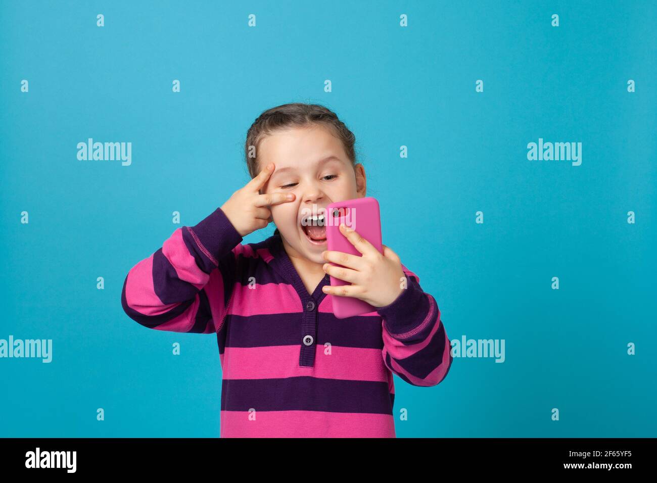 ritratto di ridente cocente ragazza con pigtail che parlano in videochiamata, prendendo selfie e mostrando segno di vittoria vicino occhio, isolato su sfondo blu Foto Stock