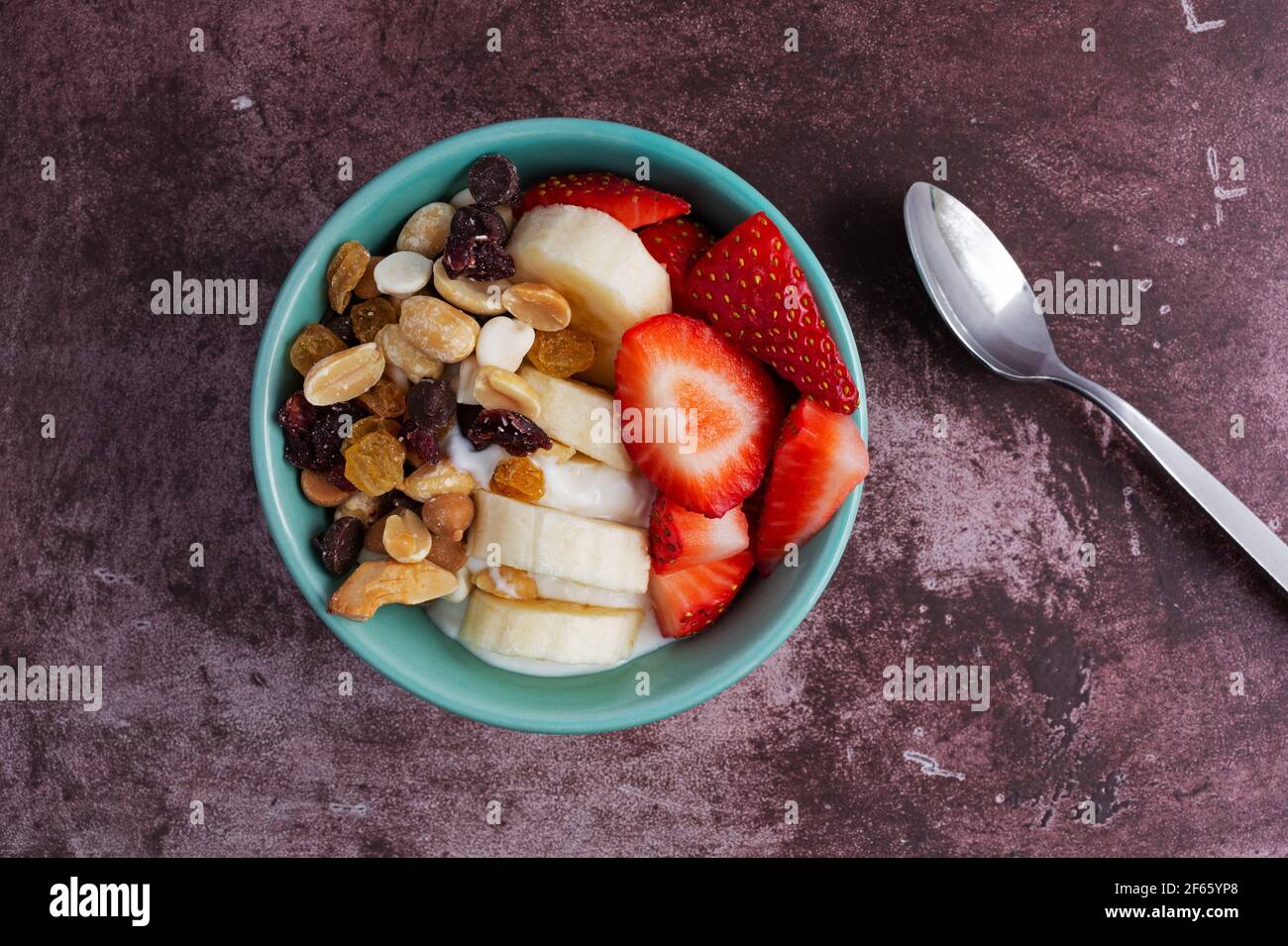 Vista dall'alto di una ciotola con pista di caramelle allo yogurt greco, mescolare banane e fragole con un cucchiaio a lato su un piano di un marronone. Foto Stock
