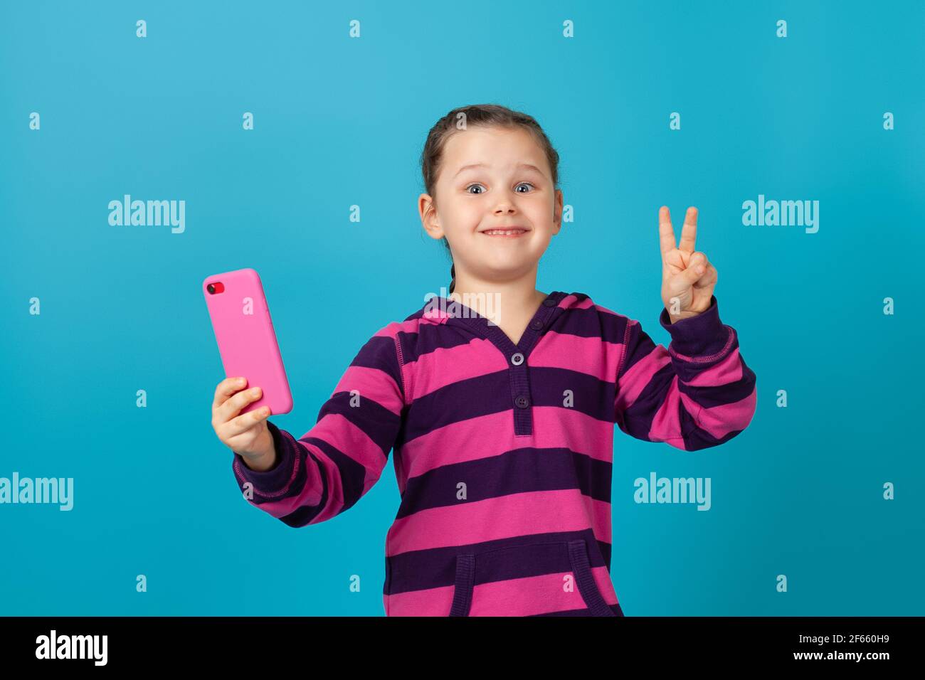 primo piano ritratto di sorpresa scioccata ragazza felice che tiene il telefono alla lunghezza del braccio, mostrando un segno di vittoria con le dita, isolato su sfondo blu Foto Stock