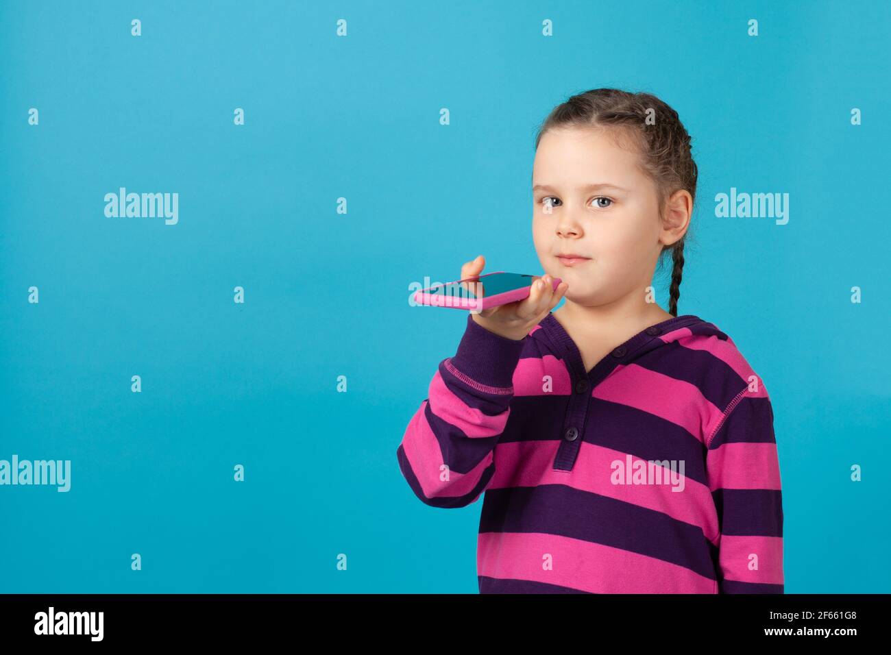 primo piano ritratto di una ragazza di cinque anni con pigtail in una felpa con cappuccio rosa a righe invia un messaggio audio sul telefono, isolato su uno sfondo blu Foto Stock
