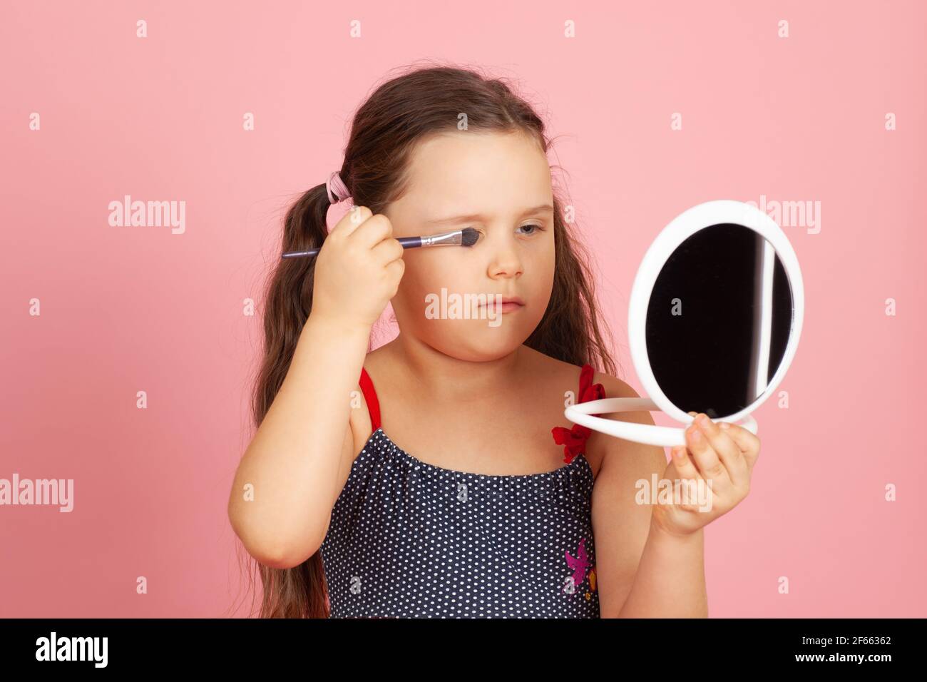 ragazza seria con i ponytails che dipingeva i suoi occhi con l'ombra degli occhi davanti ad uno specchio bianco, isolato su uno sfondo rosa Foto Stock