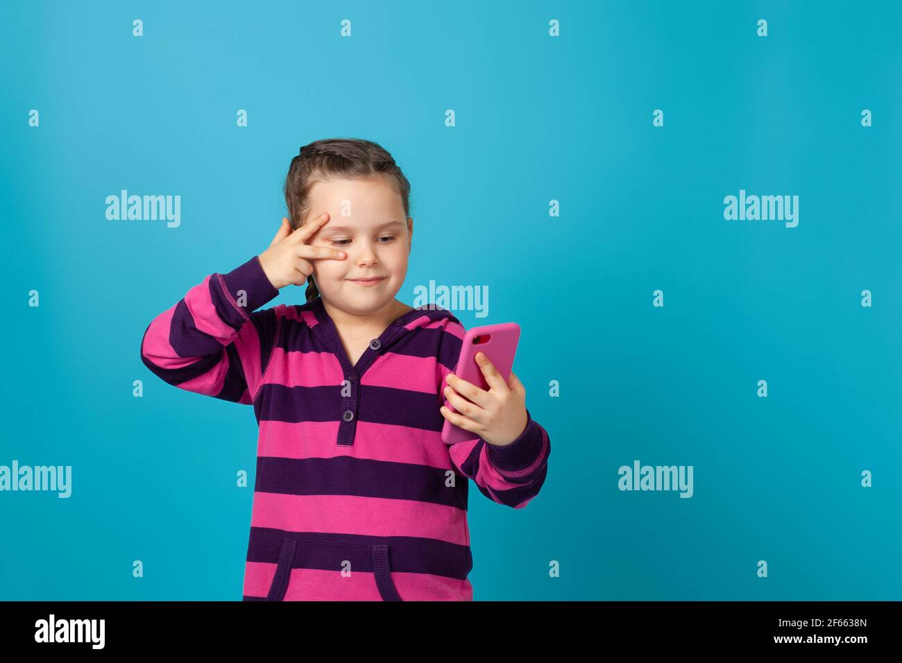 ritratto di una ragazza sorridente con i pigtail tiene in mano un telefono in un caso rosa e mostra un segno di vittoria vicino al suo occhio, isolato su uno sfondo blu Foto Stock