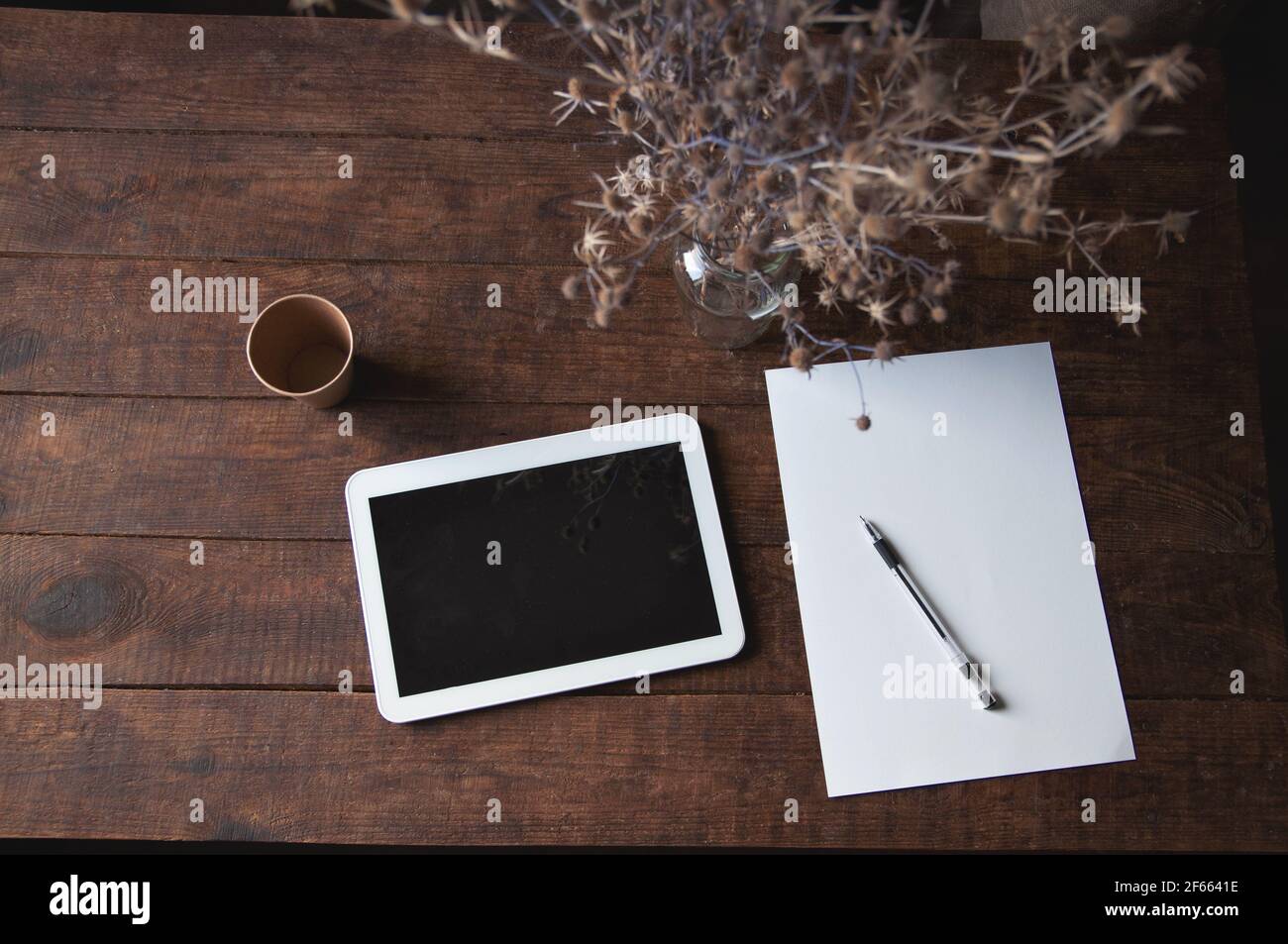 Ambiente di lavoro moderno con tablet pc sul tavolo. Sul tavolo è un  computer tablet con un foglio di carta bianca, fiori secchi in un bicchiere  e un dis Foto stock -
