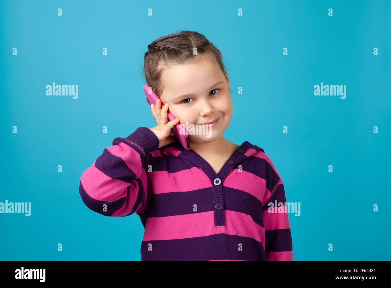 primo piano ritratto di una bella bambina sorridente con pigtail che tiene un telefono vicino all'orecchio ed effettua una telefonata, isolato su uno sfondo blu Foto Stock