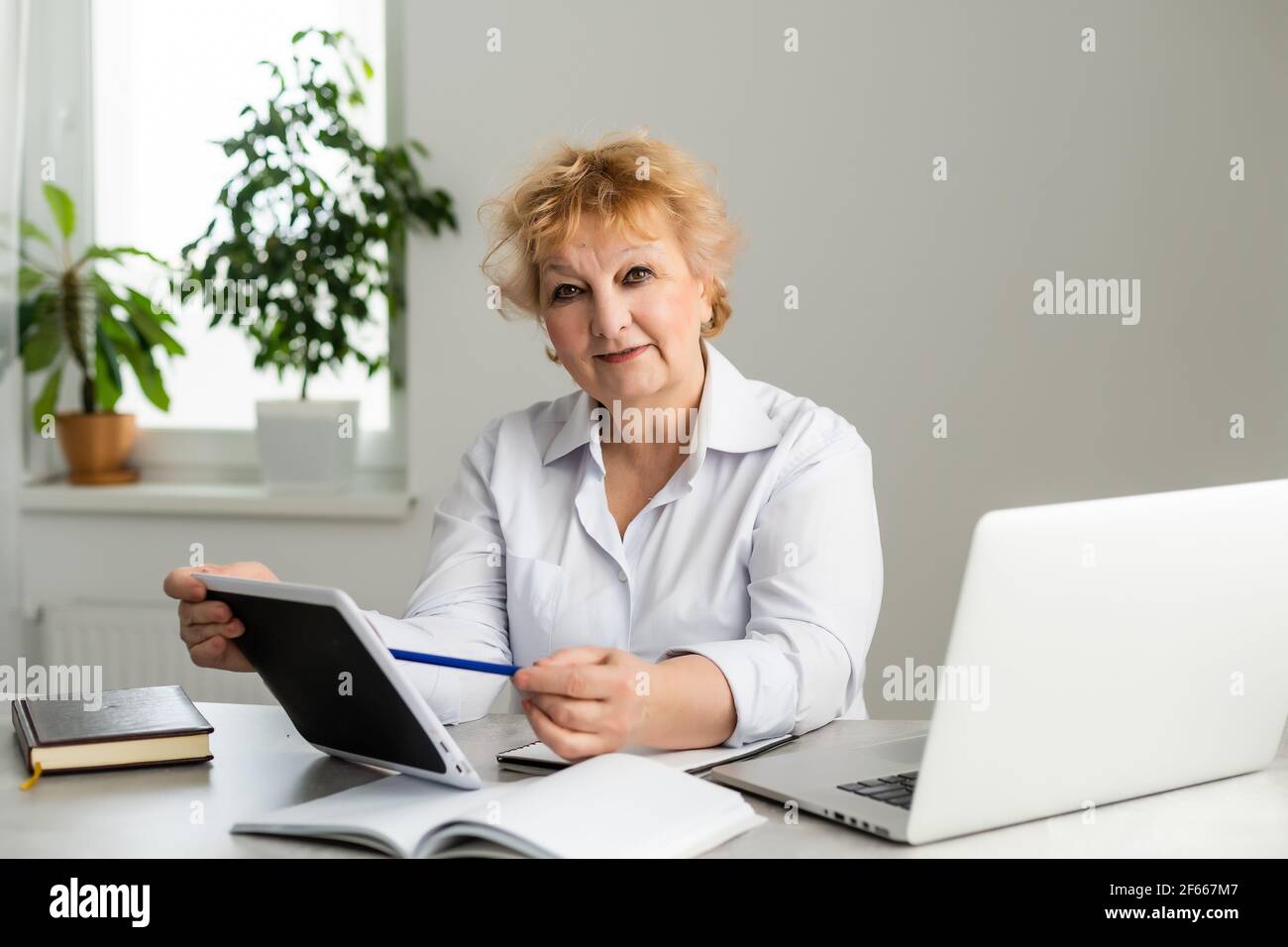 Donna anziana a casa davanti al suo computer portatile che prende appunti durante la visione di una lezione online di lingua inglese da parte di una insegnante di sesso femminile, concetto di e-learning. Foto Stock