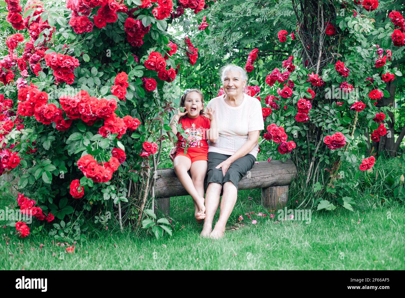 una vecchia nonna dai capelli grigi siede a piedi nudi su una panchina sotto l'arco della sposa con la nipote con lei linguetta che si aggende e gioca Foto Stock