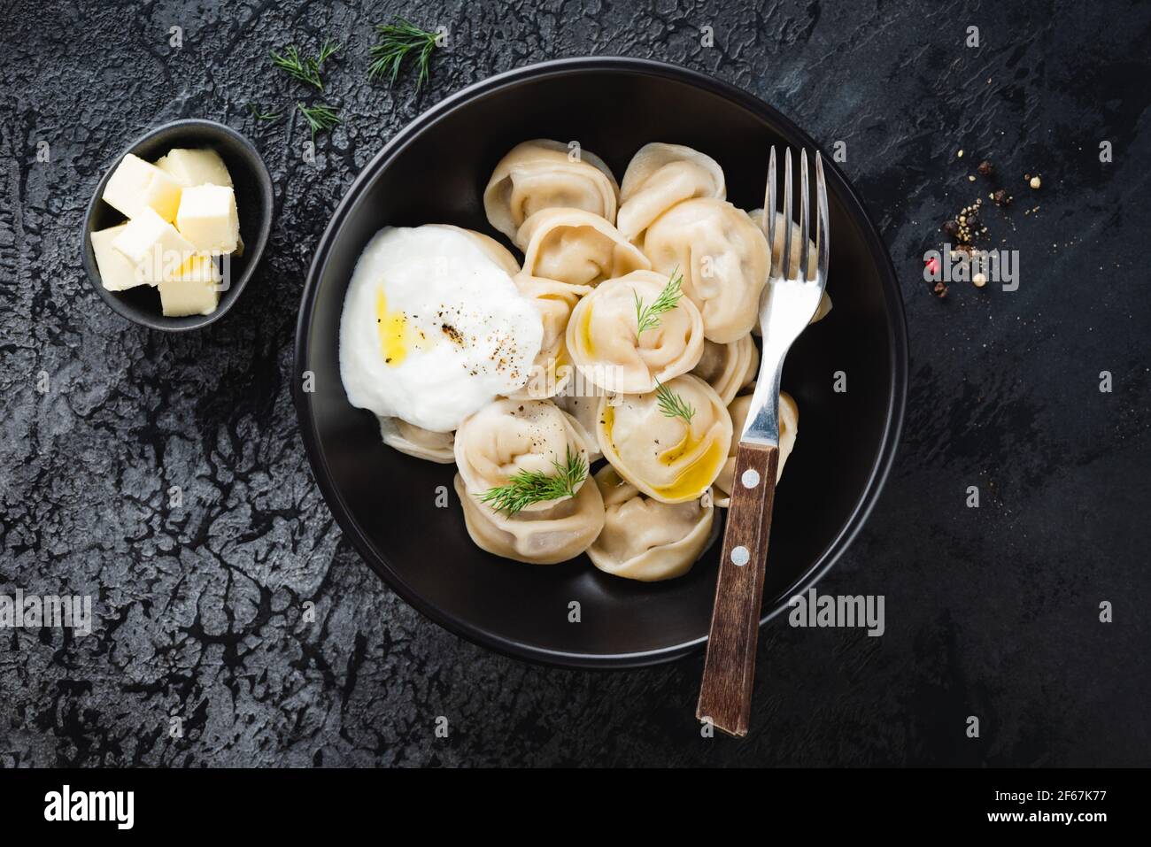 Pelmeni, gnocchi bolliti ripieni di carne con burro, aneto e panna acida su sfondo nero, vista dall'alto Foto Stock