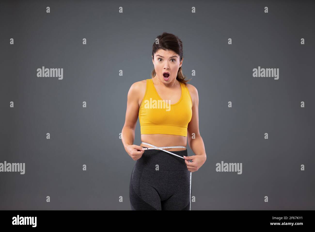 Preoccupazione per dieta sorprendentemente povera e risultati di addestramento. Una donna in abbigliamento sportivo davanti a una parete grigia che misura la vita con un nastro di misurazione. Giovane Foto Stock