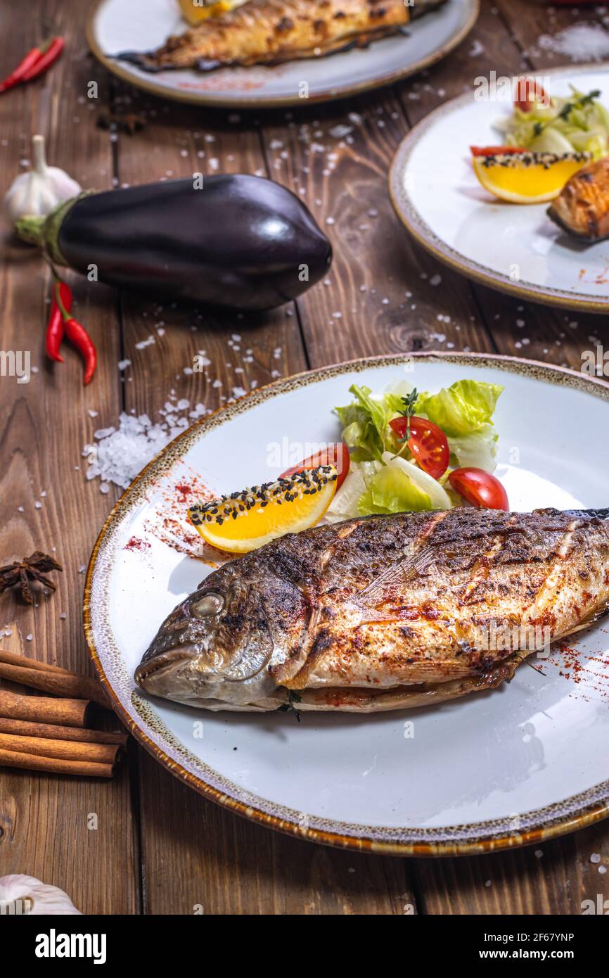 Pesce alla griglia su un piatto con verdure, peperone, sale grosso su fondo di legno marrone. Servizio ristorante. Foto Stock