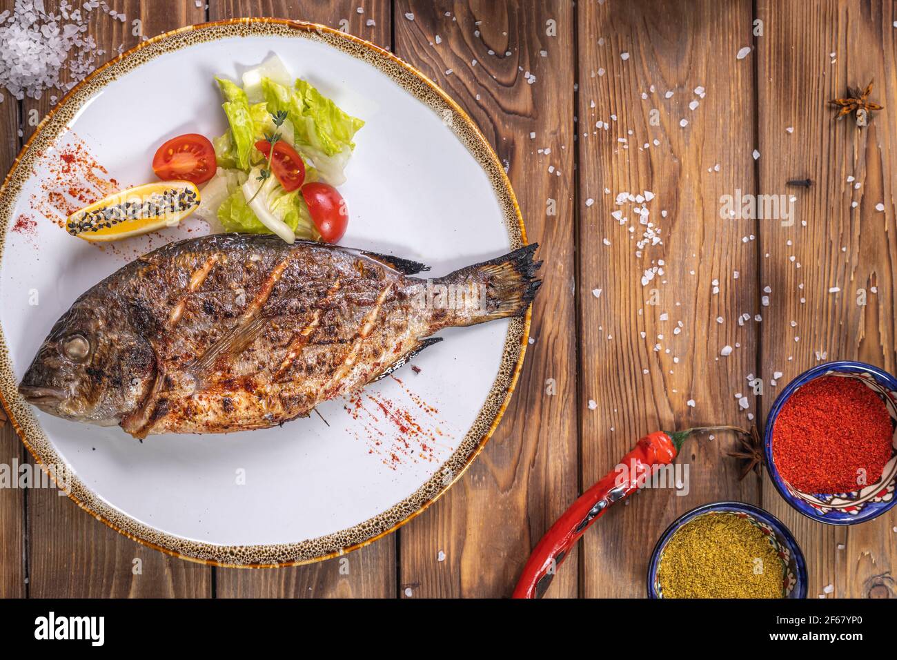Pesce alla griglia su un piatto con verdure, peperone, sale grosso su fondo di legno marrone. Servizio ristorante. Foto Stock