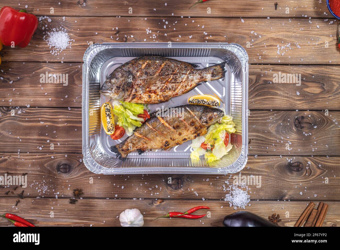 Pesce alla griglia con verdure, peperone, sale grosso, adagiato in un contenitore di alluminio su fondo di legno marrone. Consegna di cibo al ristorante. Foto Stock
