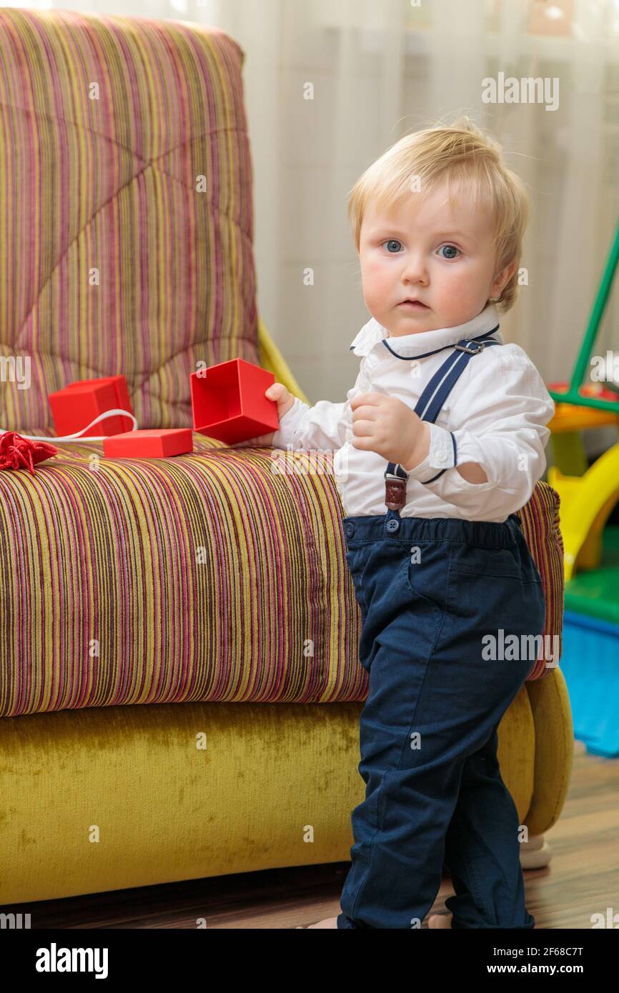 Piccolo ragazzo, bambino si alza sul divano con una scatola regalo di festa. In un bel vestito, una camicia bianca e pantaloni con sospenditori. Foto Stock