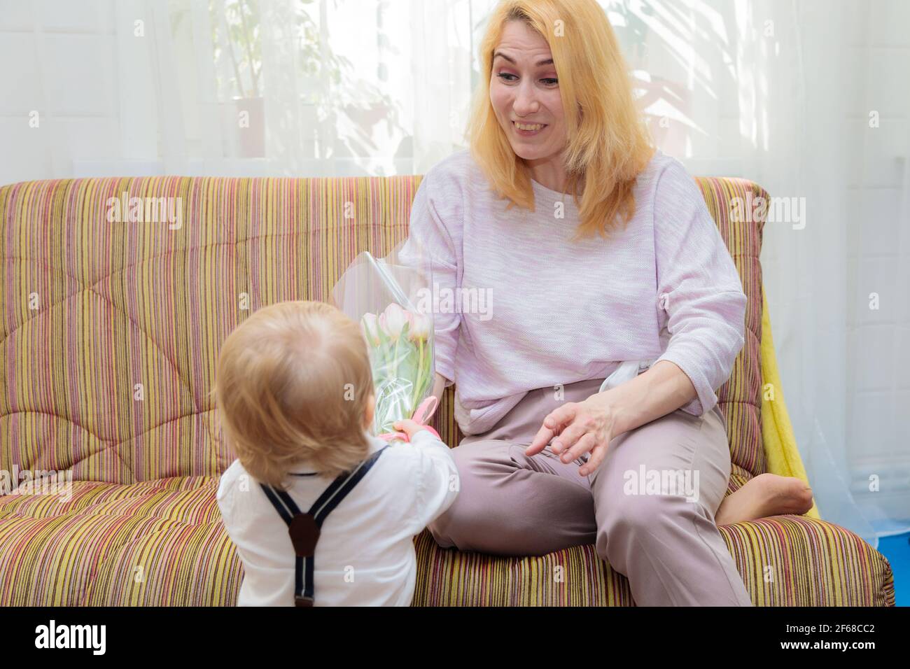 Il ragazzino si congratula con la madre per la vacanza, le dà fiori e un regalo. Ragazzo in camicia bianca e pantaloni con sospenditori Foto Stock