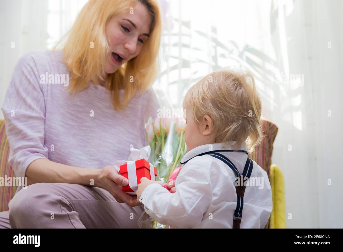 Il ragazzino si congratula con la madre per la vacanza, le dà fiori e un regalo. Ragazzo in camicia bianca e pantaloni con sospenditori Foto Stock