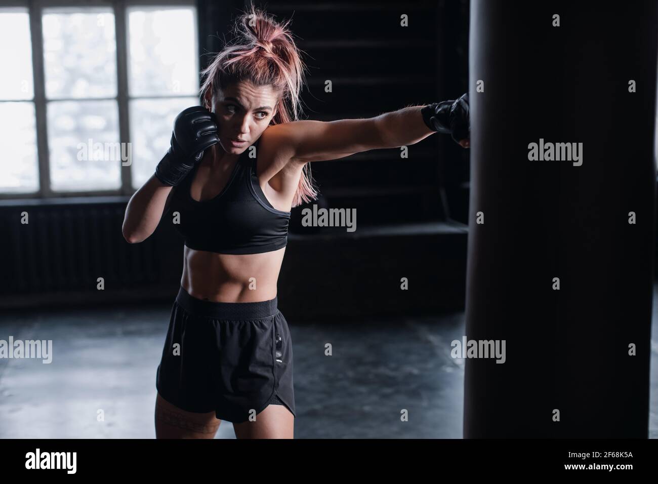 Pugile atletico FIT femmina che esercita pugni con sacchetto di boxe dentro palestra durante il kickboxing e allenamento intensivo di autodifesa Foto Stock