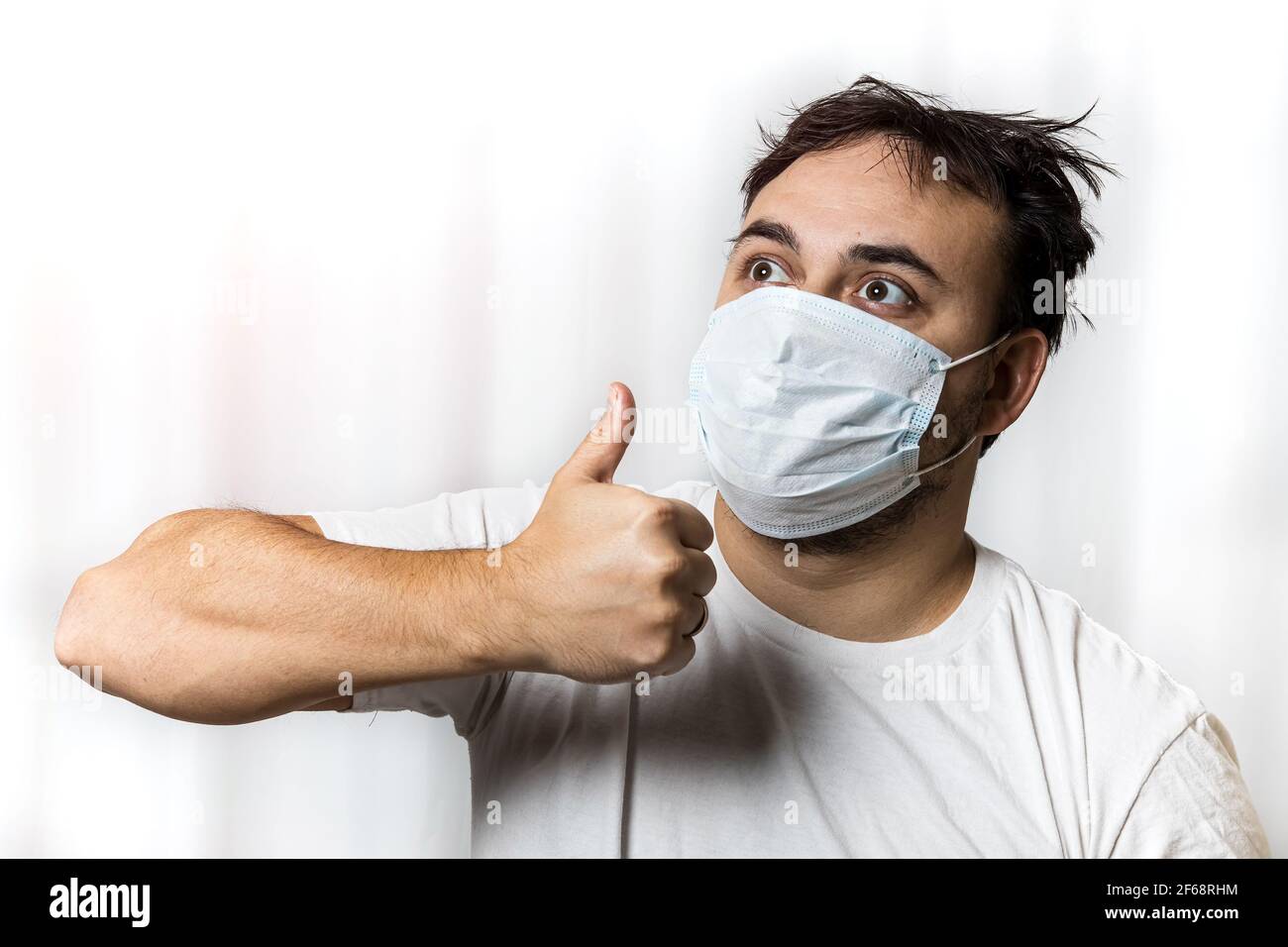 Un uomo non rasato in una maschera medica con i capelli mousled guarda a destra e in su e mostra un gesto - pollici in su. Su sfondo bianco. Foto Stock