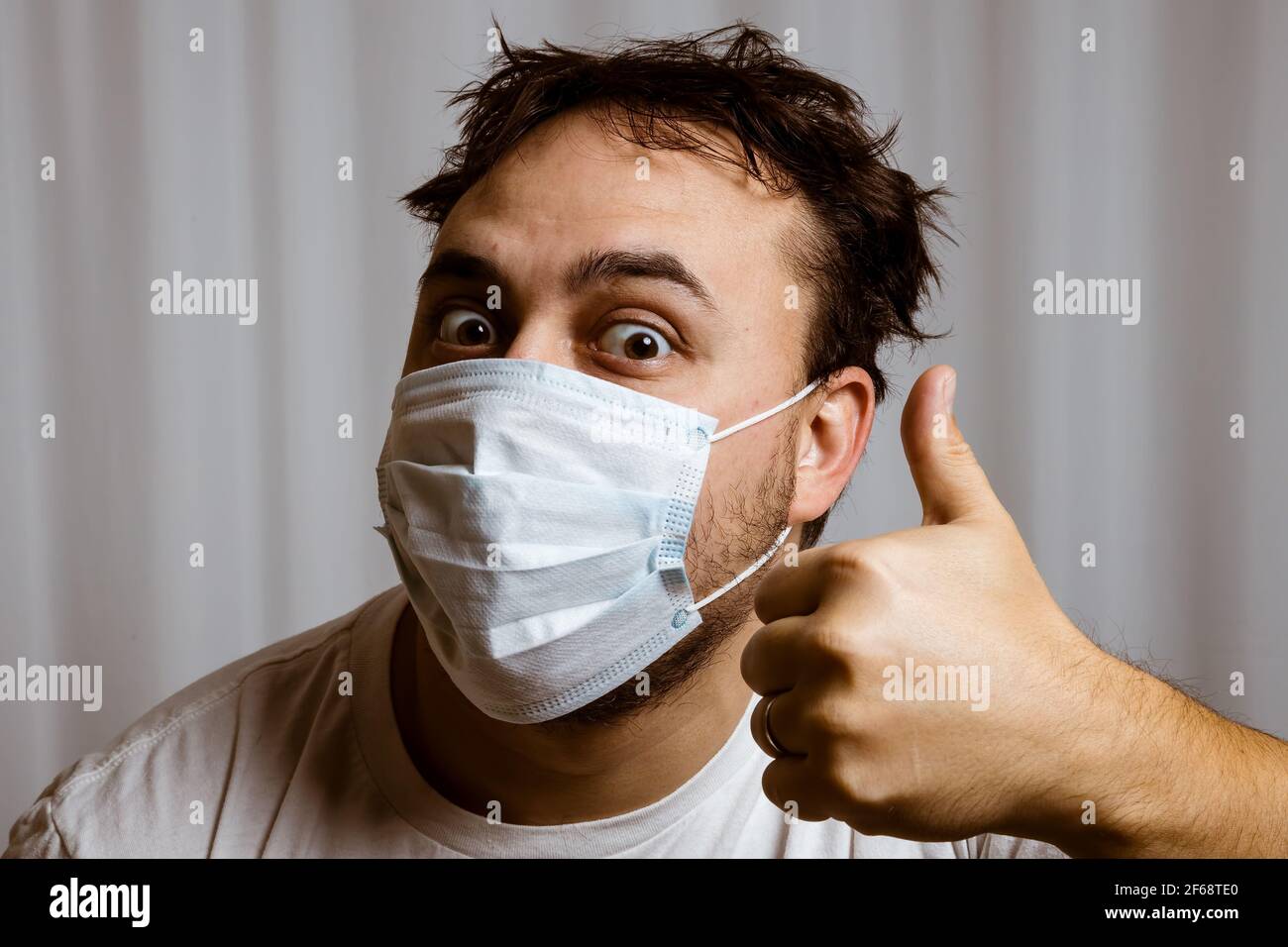 Uomo sradicato in una maschera medica bianca e t-shirt bianca con capelli ricci guarda la fotocamera e mostra un gesto - pollici verso l'alto. Su sfondo bianco. Foto Stock