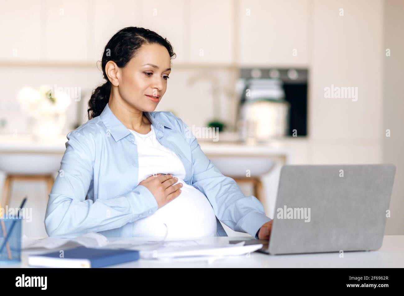 Concentrato bella corsa mista riuscita donna incinta, freelancer, manager o designer che lavorano a distanza al laptop, controllare il rapporto di affari o il progetto di progettazione, stroking ventre, sorridente Foto Stock