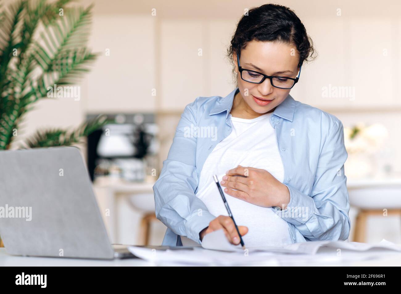 Donna di corsa mista intelligente di successo incinta con occhiali, donna di affari, broker o freelancer, lavorando a distanza, utilizza laptop, prendere appunti e controllare business plan, stracciare il ventre, sorridere Foto Stock