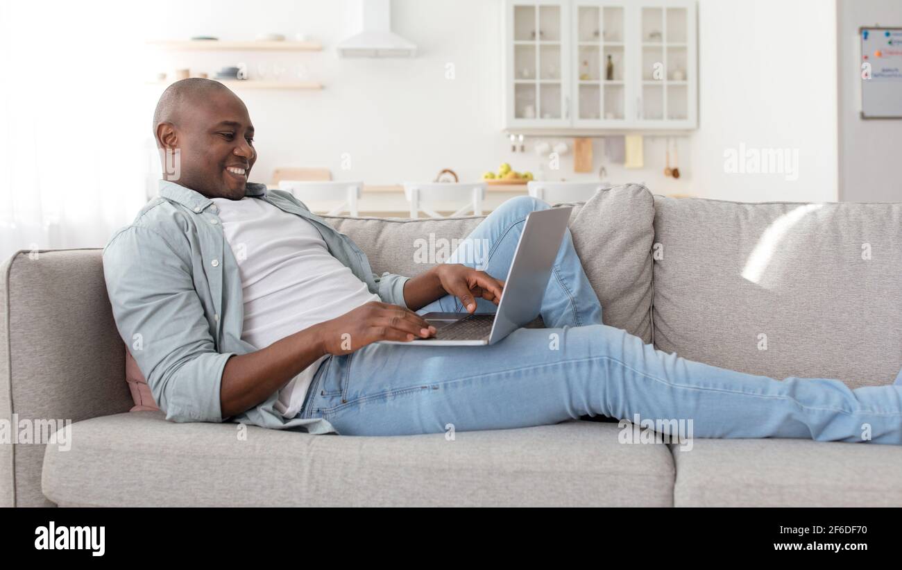 Uomo afro-americano reclinato sul divano in soggiorno, utilizzando il più recente portatile, controllare i social media, panorama Foto Stock