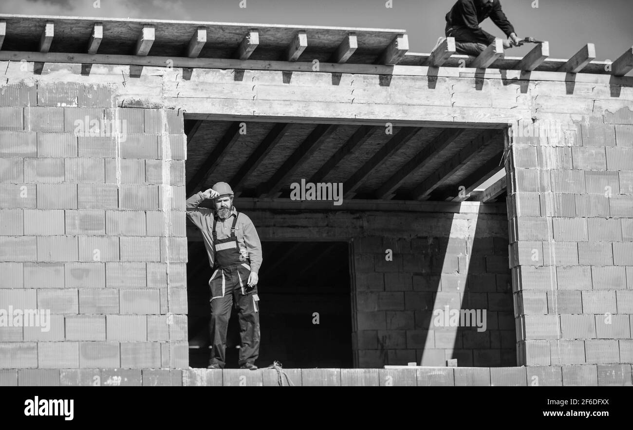 uomo bearded in uniforme e elmetto. ingegnere repairman indossare casco sul posto. costruttore stanco rilassarsi. lavoratore di costruzione fumare sigaretta. edificio è Foto Stock